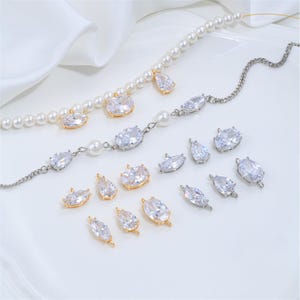 May include: A collection of jewelry components featuring white pearl strands and silver-toned chains adorned with clear, faceted, oval and teardrop-shaped cubic zirconia charms set in gold and silver metal frames, arranged on a white surface.