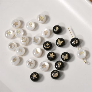 Peut inclure: Assortiment de perles rondes blanches et noires avec des accents dorés. Chaque perle présente un symbole différent, notamment des étoiles, des lunes, des papillons, des fleurs, etc.