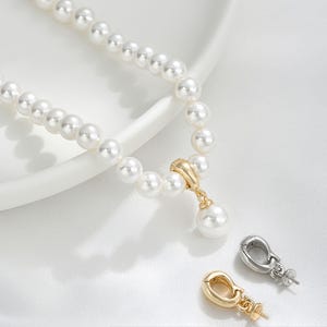May include: A white pearl necklace with a gold pendant and a single pearl drop. Two clasps, one gold and one silver, are also visible. The necklace is displayed on a white surface.