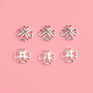 May include: Six silver-toned metal flower-shaped bead caps with a heart-shaped design. The bead caps are arranged in two rows of three on a pink background.