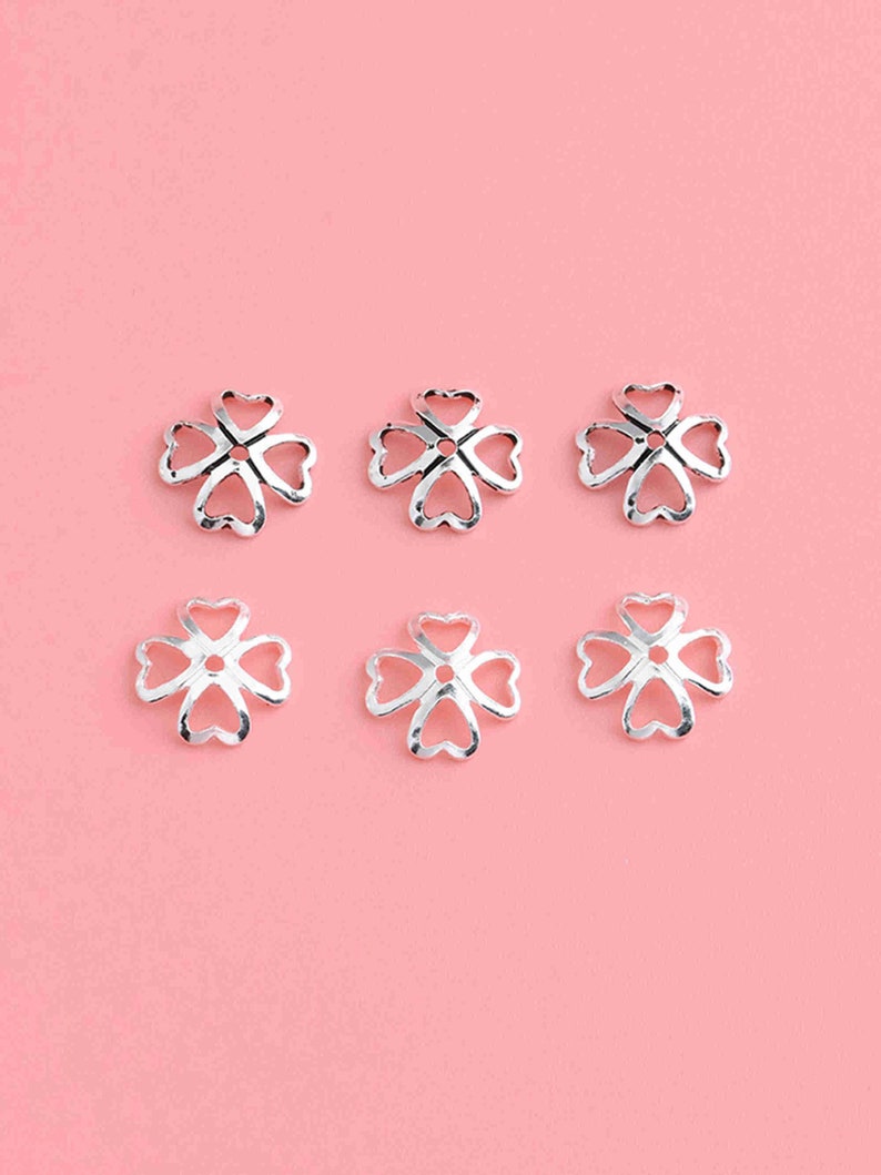 May include: Six silver-toned metal flower-shaped bead caps with a heart-shaped design. The bead caps are arranged in two rows of three on a pink background.