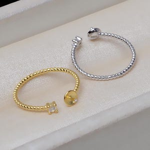 May include: Two silver rings with a textured design. One ring has a clear stone on the side and a round base for attaching a larger stone. The other ring has a clear stone on the side.