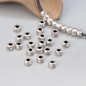 May include: A collection of silver-toned metal beads with a decorative floral pattern. The beads are round and have a hole in the center for stringing.