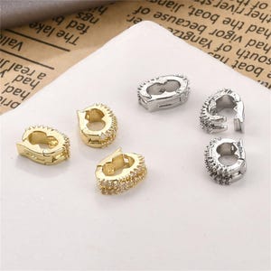 May include: Six small, decorative jewelry clasps. Three are gold-toned with clear, small stones, and three are silver-toned. The clasps are open and ready to be used. The clasps are on a white surface.