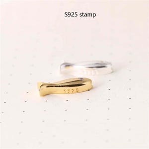 May include: A gold-coloured fish charm with the inscription "S925" on the bottom. The charm is on a white surface.
