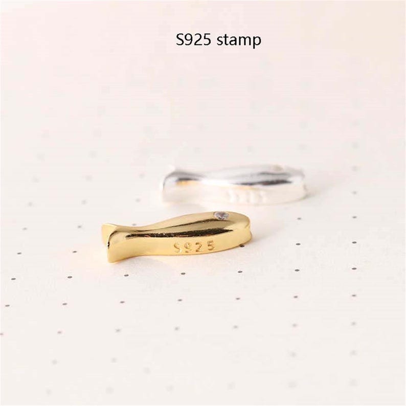 May include: A gold-coloured fish charm with the inscription "S925" on the bottom. The charm is on a white surface.