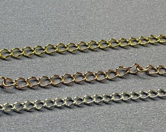 May include: Close-up of three metal chains in gold, rose gold, and silver. The chains are made of small, interlocking links, and are displayed against a gray background. The chains are likely used for jewelry making.