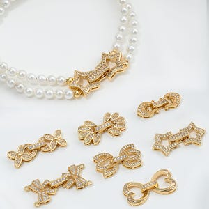 Op de afbeelding: Een set van acht gouden metalen sieraden sluitingen met heldere strass steentjes. De sluitingen zijn in verschillende vormen, waaronder sterren, strikken en harten.