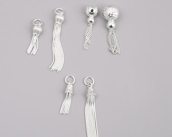 May include: A collection of silver-colored tassel charms in various designs. Some have beaded tops, while others feature intricate chain or strand details. The charms are displayed against a neutral gray background, showcasing their reflective surfaces.