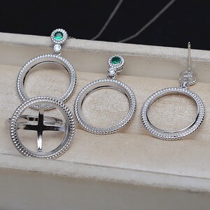 Puede incluir: Un conjunto de collar, anillo y pendientes de plata con un engaste redondo para una moneda o un amuleto. El conjunto presenta un acento de piedra preciosa verde.