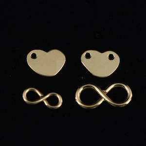 Peut inclure: Quatre apprêts de bijoux dorés sur fond noir. Deux sont en forme de cœur avec des trous, et deux sont des symboles de l'infini. Ces apprêts sont probablement utilisés pour la fabrication de bijoux, tels que des boucles d'oreilles ou des pendentifs.