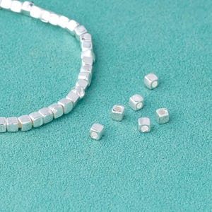 Può includere: Un braccialetto d'argento fatto di piccole perline quadrate. Il braccialetto è su una superficie turchese con diverse perline quadrate d'argento sciolte sparse intorno.