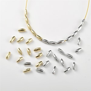 May include: A collection of gold and silver oval-shaped beads. The beads are strung on a wire necklace. The beads are scattered on a white surface.