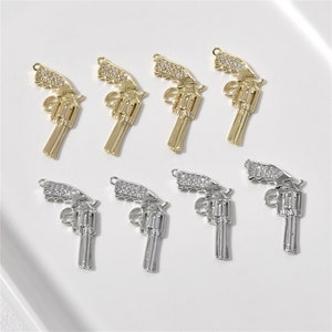 May include: A set of eight silver and gold charm pendants in the shape of a revolver pistol. Each charm is decorated with clear rhinestones.