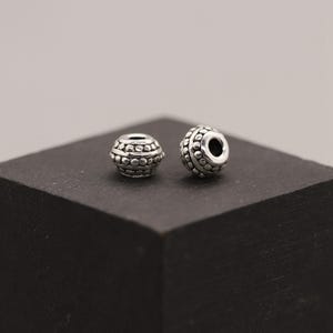 Op de afbeelding: Twee kleine, zilverkleurige kralen met een gestructureerd, gestippeld patroon. Elke kraal heeft een centraal gat en een reeks verhoogde stippen die de kraal omcirkelen. De kralen worden gepresenteerd op een donker, vierkant oppervlak.