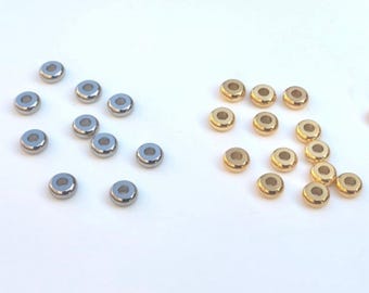Könnte beinhalten: Eine Sammlung kleiner Metallperlen in Silber und Gold. Die runden Perlen haben ein Loch in der Mitte und eignen sich für Schmuckherstellung oder Bastelarbeiten. Die Perlen sind auf einer weißen Oberfläche verstreut.