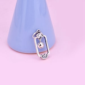 May include: A silver charm featuring a square frame with a dangling ball. The charm is intricately detailed and has a delicate design.