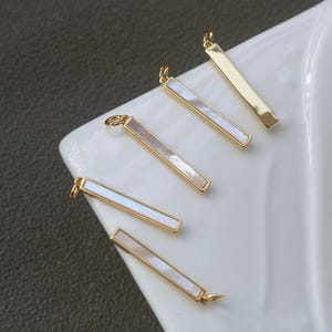 May include: Gold-tone bar pendants with a rectangular mother-of-pearl inlay. The pendants have a small loop at the top for attaching to jewelry. The pendants are displayed on a white surface.