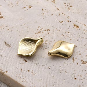 Könnte beinhalten: Zwei goldfarbene Metallperlen in Form stilisierter Blütenblätter. Eine Perle weist eine texturierte, gestreifte Oberfläche auf, während die andere eine glatte, polierte und reflektierende Oberfläche hat. Jede Perle hat ein Loch am Ende.