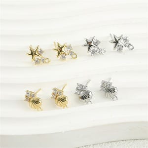 Op de afbeelding: Een assortiment oorstekers in goud- en zilverkleuren. Ontwerpen omvatten ster- en schelpvormen, elk versierd met kleine, sprankelende kristallen. De oorbellen worden gepresenteerd op een witte ondergrond.