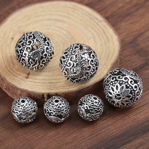 May include: Six silver-toned metal beads with intricate, ornate designs. The beads are round and have a hollow center. The beads are arranged in a semi-circle on a wooden surface.