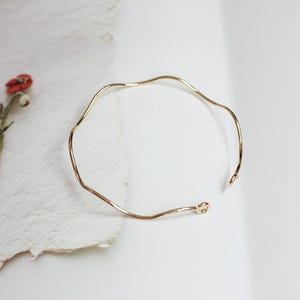 Op de afbeelding: Een goudkleurige, open manchetarmband met een golvend, organisch ontwerp. De armband heeft een gladde, gepolijste afwerking en een delicate, minimalistische esthetiek. De open uiteinden van de armband zijn licht afgerond.