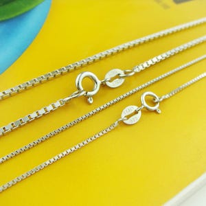May include: Close-up of several silver box chain necklaces with lobster claw clasps. The chains vary in thickness and are displayed against a bright yellow background. Each chain has a small tag near the clasp.