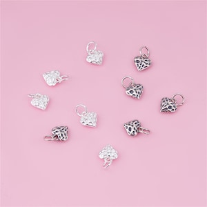 May include: A collection of ten silver heart-shaped charms with a textured surface. The charms are scattered on a pink background.