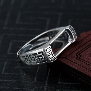 May include: A silver-colored ring with a geometric design. The ring features a rectangular face with a maze-like pattern and a smooth, rounded band. The ring is displayed on a dark surface.