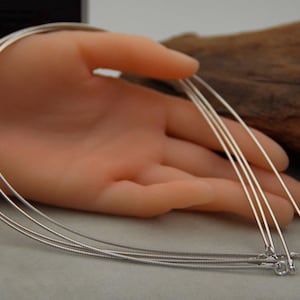 Pode incluir: Quatro colares de corrente de prata com fechos de lagosta. As correntes estão dispostas sobre uma mão e uma superfície de madeira.