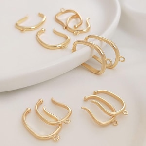 May include: A collection of gold-colored jewelry findings, including curved and rectangular shapes. Each piece has a small loop for attaching to other components. The findings are arranged on a white surface.