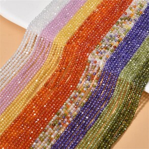 Könnte beinhalten: Sortiment an facettierten Edelsteinperlen in verschiedenen Farben, darunter klar, Lavendel, Gelb, Orange, Rot, Blau, Grün und Braun. Die Perlen sind auf einem weißen Hintergrund aufgereiht.
