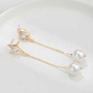 Pode incluir: Um par de brincos pendurados dourados. Cada brinco apresenta um topo em forma de estrela com uma pedra preciosa transparente, uma corrente delicada e uma grande pérola branca redonda na parte inferior. Os brincos estão em uma superfície branca.