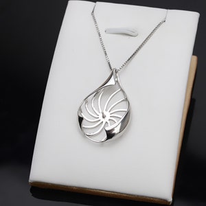 Puede incluir: Un collar de plata con un colgante en forma de concha de nautilus. El colgante está abierto y tiene un diseño en espiral.