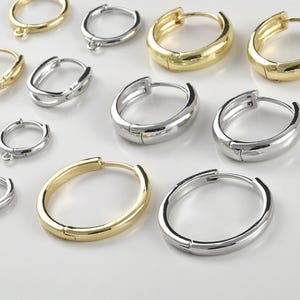 Pode incluir: Uma variedade de brincos de argola dourados e prateados. Os brincos variam em tamanho e design, alguns com fecho articulado. São exibidos em uma superfície branca, mostrando seu acabamento polido.