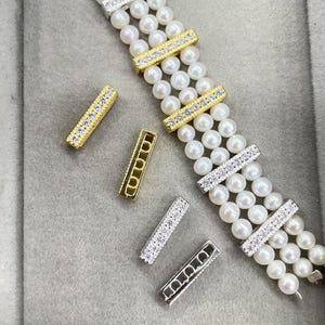 May include: A multi-strand pearl bracelet featuring three rows of small white spherical pearls. The bracelet is accented with rectangular gold and silver-toned metal spacers embedded with clear crystals. Four additional loose rectangular spacers are shown.