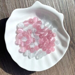 May include: A white bowl filled with pink and white round beads. The beads are smooth and have a slight shine.