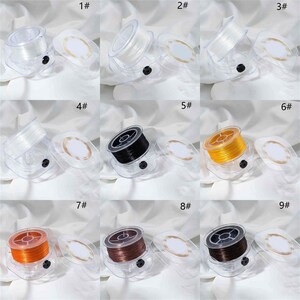 Op de afbeelding: Negen spoelen van helder plastic garen, elk met een anders gekleurd garen eromheen gewikkeld. De kleuren zijn helder, wit, zwart, oranje, goud, bruin en donkeroranje. De spoelen zijn genummerd van 1 tot en met 9.