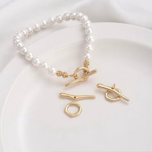Pode incluir: Uma pulseira de pérolas brancas com fecho de alternância dourado. A pulseira está sobre uma superfície branca, com três fechos de alternância dourados adicionais. Uma peça de joalharia clássica.