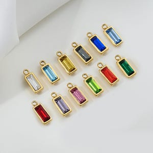 Puede incluir: Una colección de colgantes rectangulares de piedras preciosas en varios colores, incluyendo azul, verde, rojo y amarillo. Cada colgante tiene un marco dorado y un pequeño lazo en la parte superior para sujetarlo a la joyería. Las piedras preciosas están dispuestas sobre una superficie blanca.