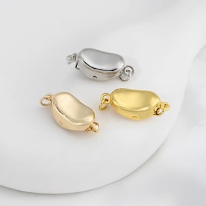 Può includere: Tre chiusure per gioielli in metallo nei toni dell'oro e dell'argento. Le chiusure hanno una forma arrotondata a fagiolo con piccoli anelli su ciascuna estremità per il fissaggio ai gioielli. Le chiusure sono su una superficie bianca.