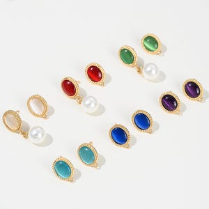 Puede incluir: Una colección de pendientes de piedras preciosas de colores. Cada pendiente presenta una piedra de forma ovalada en varios colores, como rojo, verde, azul y morado, engastada en un marco dorado. Algunos pendientes tienen una perla blanca colgante.