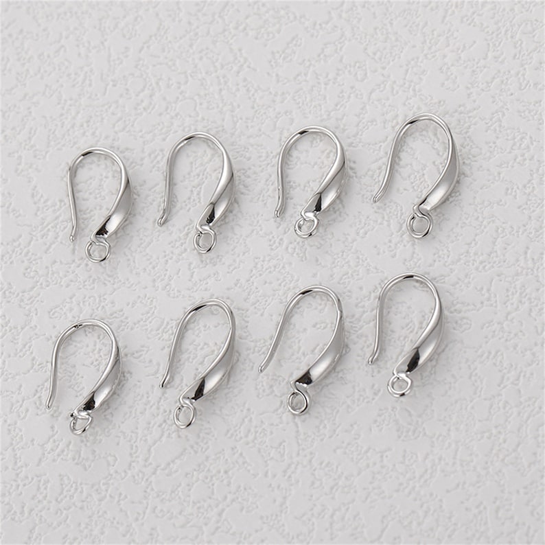 May include: Eight silver-colored earring hooks. Each hook has a curved design with a loop at the bottom for attaching beads or charms. The hooks are displayed on a textured, off-white surface.