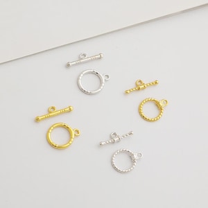 May include: A selection of gold and silver-toned jewellery clasps. The image shows toggle bars and circular rings, with different textures and finishes. These clasps are designed for jewellery making and crafting.