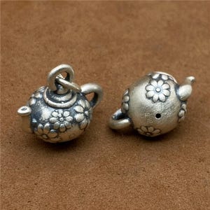 Puede incluir: Dos pequeños colgantes de tetera plateados. Cada colgante está decorado con un motivo floral y tiene un asa, un pico y un lazo para sujetarlo a una cadena o pulsera. Los colgantes se muestran sobre una superficie marrón.
