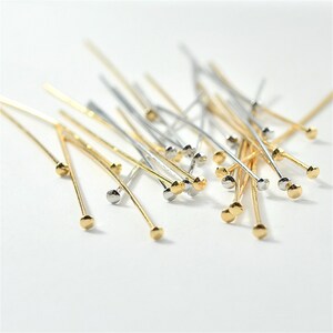 Puede incluir: Una colecci&oacute;n de alfileres de cabeza redonda de oro y plata. Los alfileres est&aacute;n esparcidos sobre una superficie blanca.