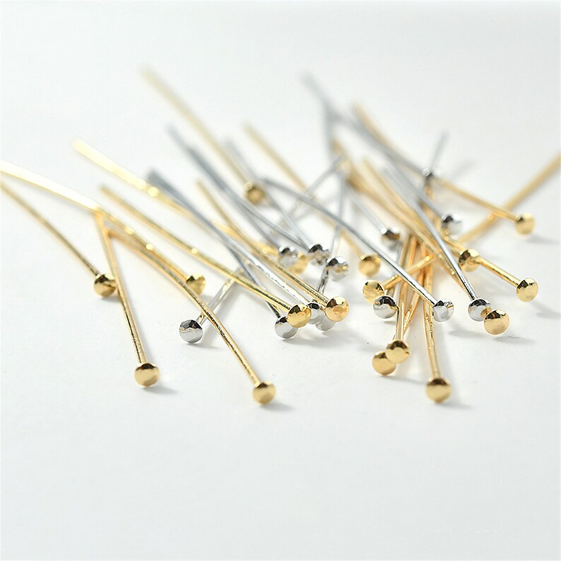 Puede incluir: Una colecci&oacute;n de alfileres de cabeza redonda de oro y plata. Los alfileres est&aacute;n esparcidos sobre una superficie blanca.