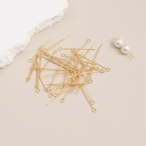 Peut inclure: Un ensemble d'épingles à tête dorées et deux perles blanches en fausse perle sur une surface beige clair. Les épingles à tête ont une boucle à une extrémité et une épingle droite à l'autre. Les perles sont fixées à une épingle dorée.