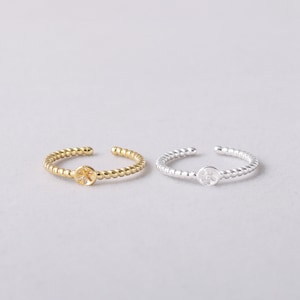 May include: Two dainty adjustable rings with a twisted beaded band and a small four-leaf clover charm. The ring on the left is gold and the ring on the right is silver.