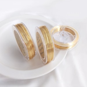 Könnte beinhalten: Drei Spulen goldfarbenen Drahtes auf einem weißen Teller. Jede Spule ist mit "0.4mm Jewelry Wire" beschriftet.